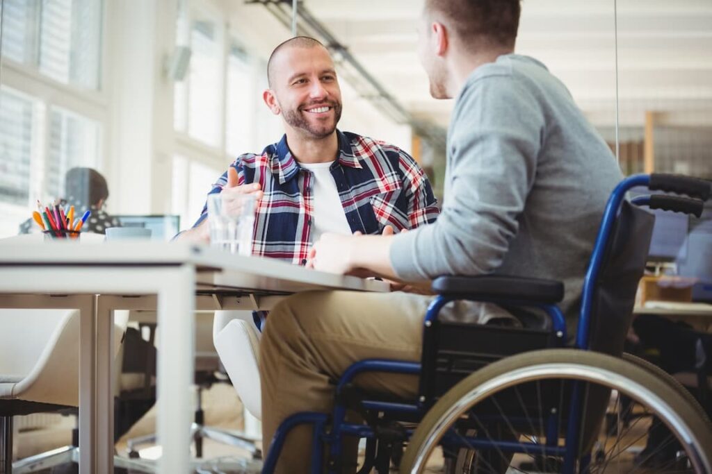 Two men in wheelchairs talking at a table in a home office.